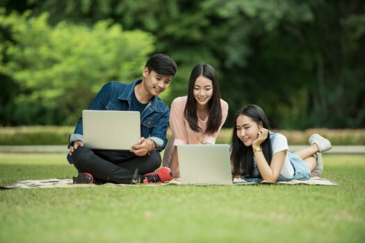 tiga mahasiswa sedang belajar bersama di sebuah taman dengan membawa laptop